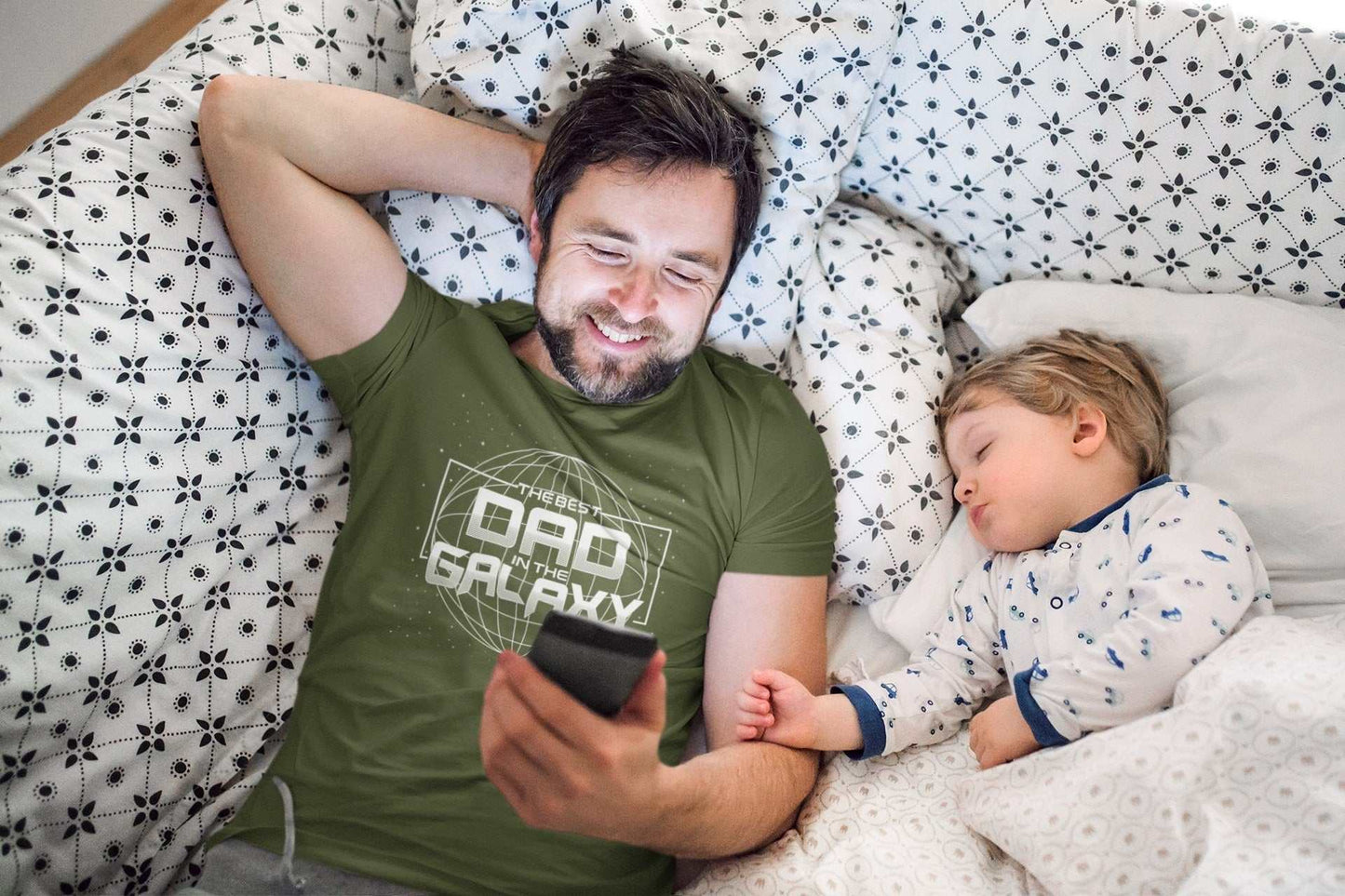 Dad wearing "Best Dad in the Galaxy" shirt on bed with sleeping child, Father's Day gift.