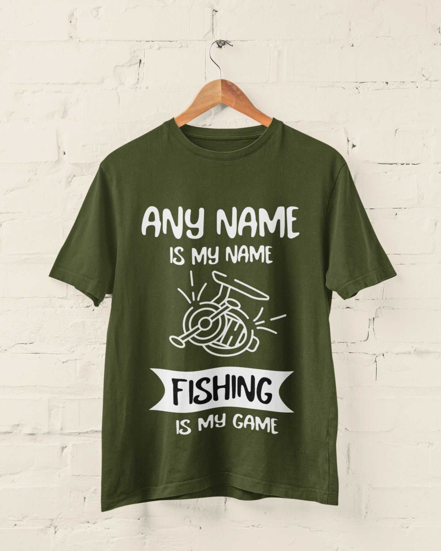 Personalised fishing T-shirt with "Any Name is My Name, Fishing is My Game" design hanging on a white brick wall.