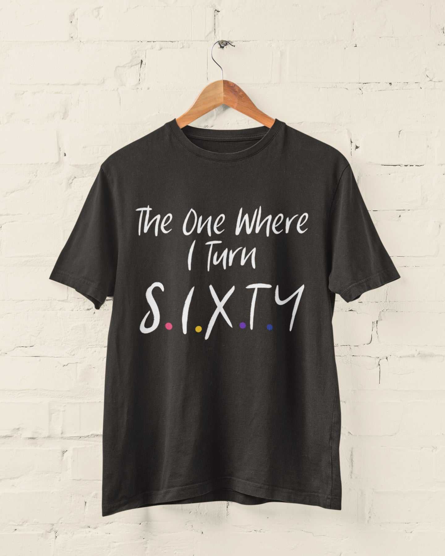 Funny 60th birthday t-shirt with Friends TV show font, "The One Where I Turn Sixty", hanging on a wooden hanger against a brick wall.