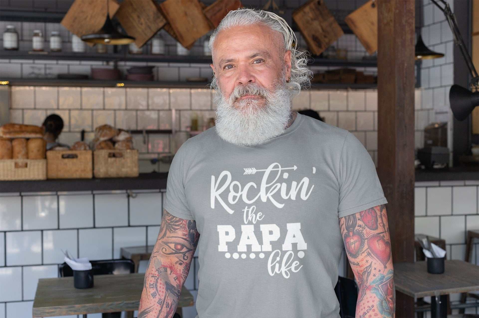 "Rockin' the Papa Life" t-shirt modeled by a bearded man in a rustic bakery setting.
