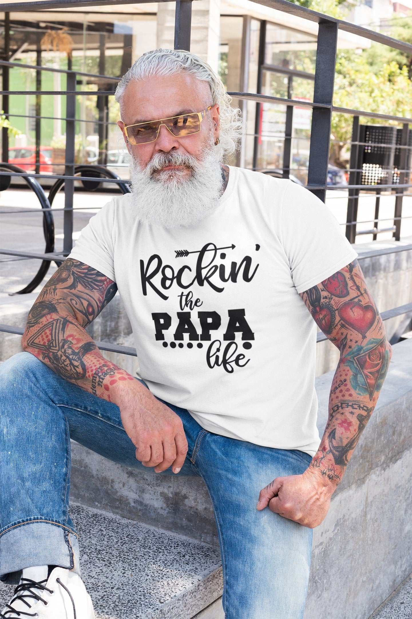Stylish "Rockin' the Papa Life" t-shirt worn by a tattooed man sitting outdoors, ideal gift for papas.